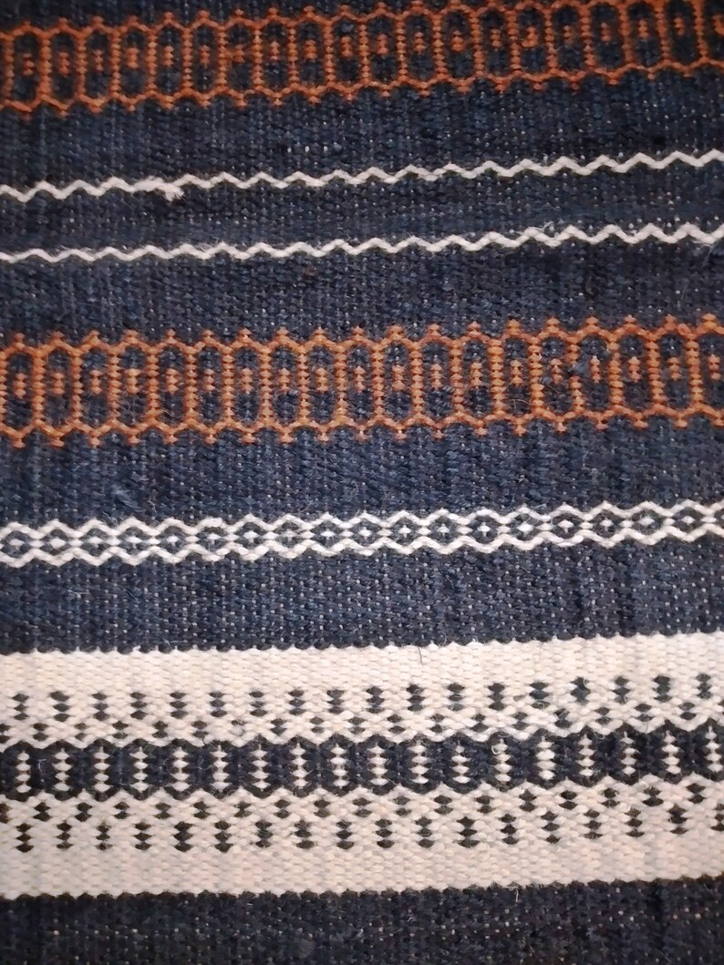 May include: A close-up of a woven fabric with a repeating pattern of brown, white, and black geometric shapes. The fabric has a textured surface and a dark blue background.