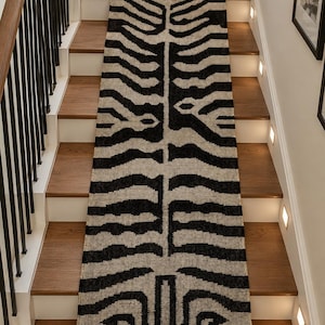 Pode incluir: Um tapete passadeira longo e retangular com um padrão animal de alto contraste inspirado na zebra, em preto e bege. O tecido apresenta um design simétrico ao longo do centro e é rematado com franjas curtas e escuras em ambas as extremidades.