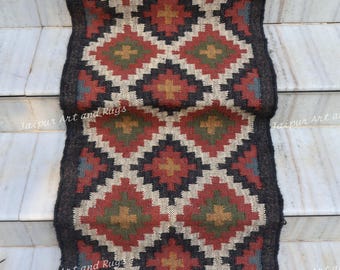 May include: A rectangular rug with a geometric diamond pattern in red, black, cream, green, and gold. The rug has a dark gray border and fringe on the bottom. The words "Jaipur Art and Rugs" are printed on the rug.