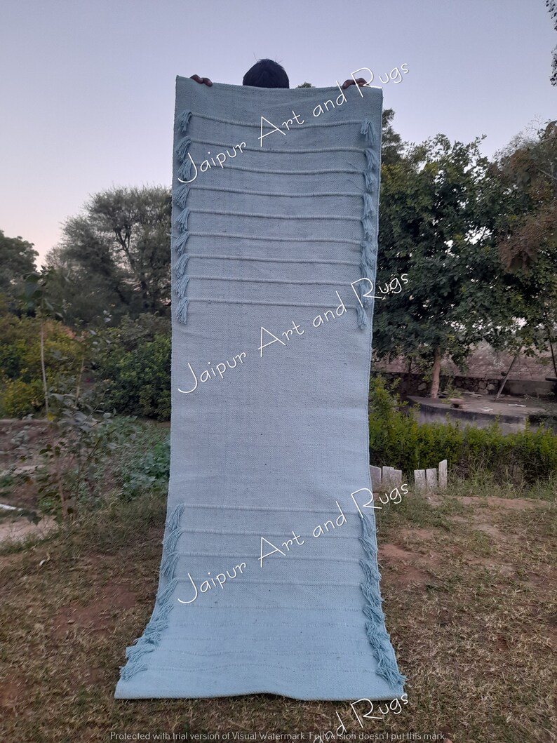 May include: A light blue handwoven rug with fringe on both ends. The rug is held up by a person, and the text "Jaipur Art and Rugs" is visible on the rug.