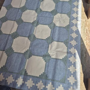 May include: A rectangular area rug featuring a repeating geometric pattern of light cream squares set against a textured blue background. The design includes a decorative border with a repeating cross-like motif in cream, framing the central grid.