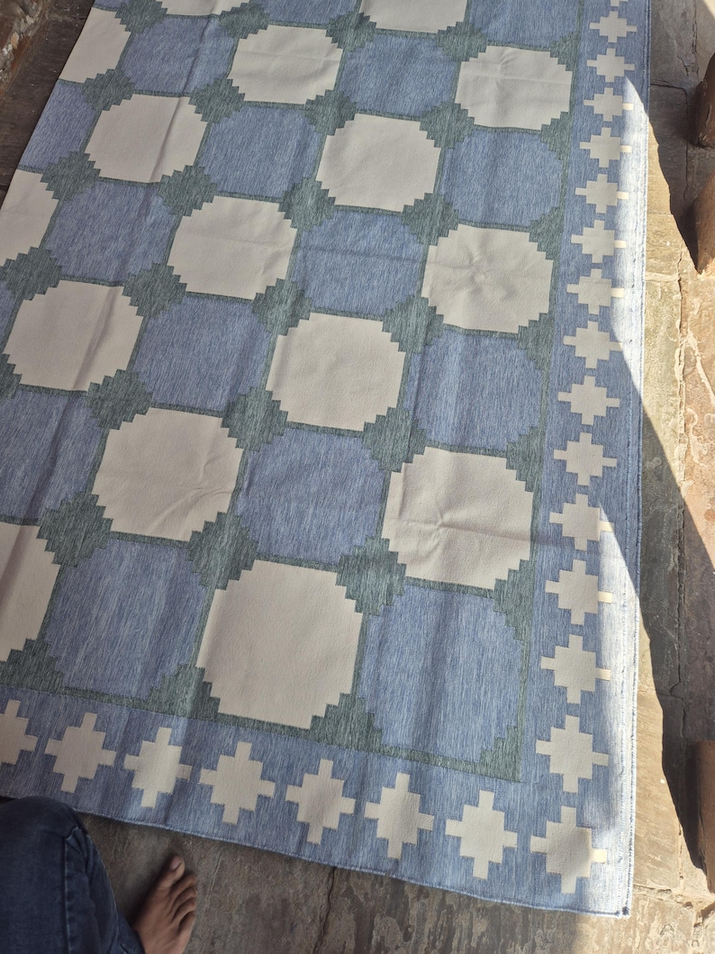 May include: A rectangular area rug featuring a repeating geometric pattern of light cream squares set against a textured blue background. The design includes a decorative border with a repeating cross-like motif in cream, framing the central grid.