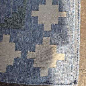 May include: A woven area rug featuring a repeating geometric cross pattern in off-white against a textured medium blue background. The rug has a finished edge with a visible stitched border and a dense, flat-weave construction throughout the surface.