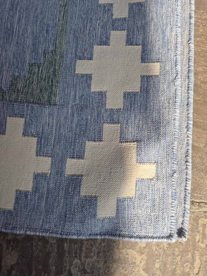 May include: A woven area rug featuring a repeating geometric cross pattern in off-white against a textured medium blue background. The rug has a finished edge with a visible stitched border and a dense, flat-weave construction throughout the surface.