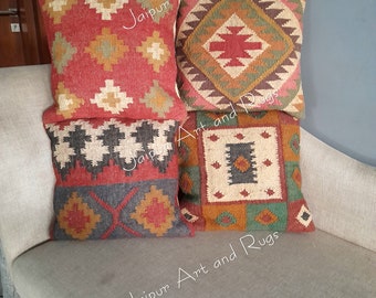 May include: Four decorative throw pillows with geometric patterns in shades of red, orange, brown, green, and black. The pillows are made of woven fabric and have a rustic, bohemian style. The pillows are arranged on a light gray couch. The text "Jaipur Art and Rugs" is visible on the pillows.