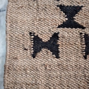 May include: Close-up of a jute rug with a black and white geometric pattern. The rug has a natural, earthy texture and is woven with a simple, repeating design.