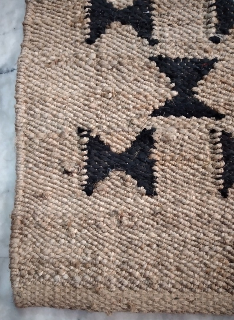 May include: Close-up of a jute rug with a black and white geometric pattern. The rug has a natural, earthy texture and is woven with a simple, repeating design.