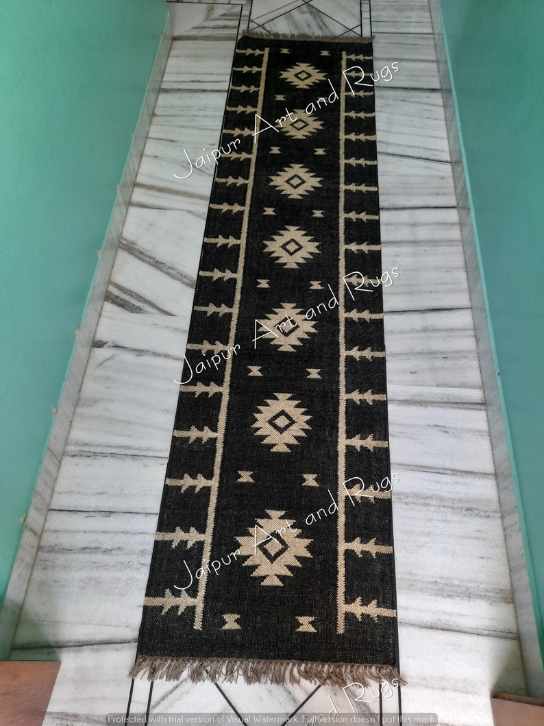 May include: Black and cream geometric patterned rug with fringe on the ends. The rug is laid out on a white marble floor.