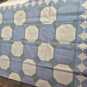 May include: A rectangular area rug featuring a repeating geometric pattern of large cream-colored octagons set against a blue background. The design includes a border with a repeating cross motif in cream, with dark green accents at the corners.