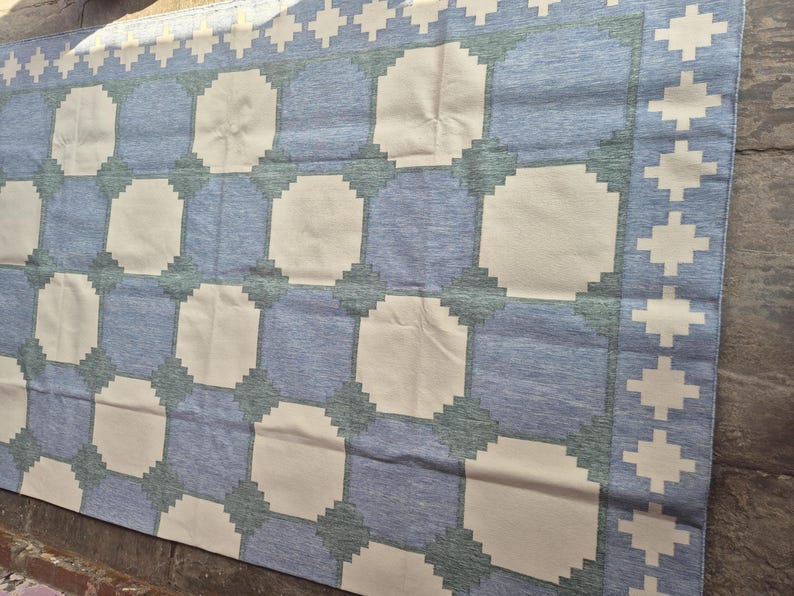 May include: A rectangular area rug featuring a repeating geometric pattern of large cream-colored octagons set against a blue background. The design includes a border with a repeating cross motif in cream, with dark green accents at the corners.