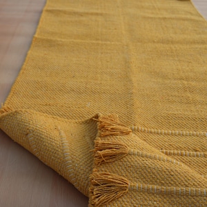 Op de afbeelding: Een lange, rechthoekige mosterdgele loper met een geweven textuur, horizontale witte stiksels en decoratieve kwastjes aan de korte uiteinden. Het kleed is in een hoek omgeslagen, waardoor de onderkant en de weefstructuur zichtbaar zijn.