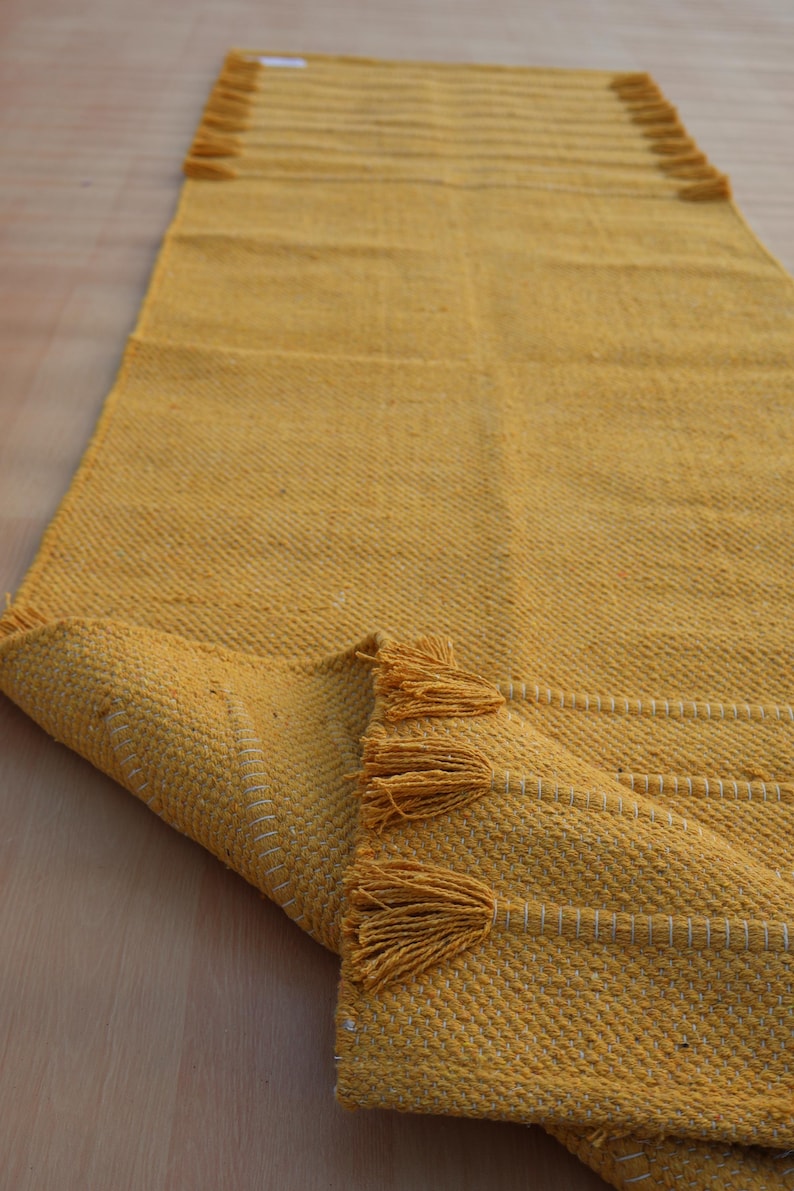 Op de afbeelding: Een lange, rechthoekige mosterdgele loper met een geweven textuur, horizontale witte stiksels en decoratieve kwastjes aan de korte uiteinden. Het kleed is in een hoek omgeslagen, waardoor de onderkant en de weefstructuur zichtbaar zijn.