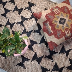 May include: A black and beige jute rug with a geometric pattern. A red, yellow, and gray patterned pillow is on top of the rug.