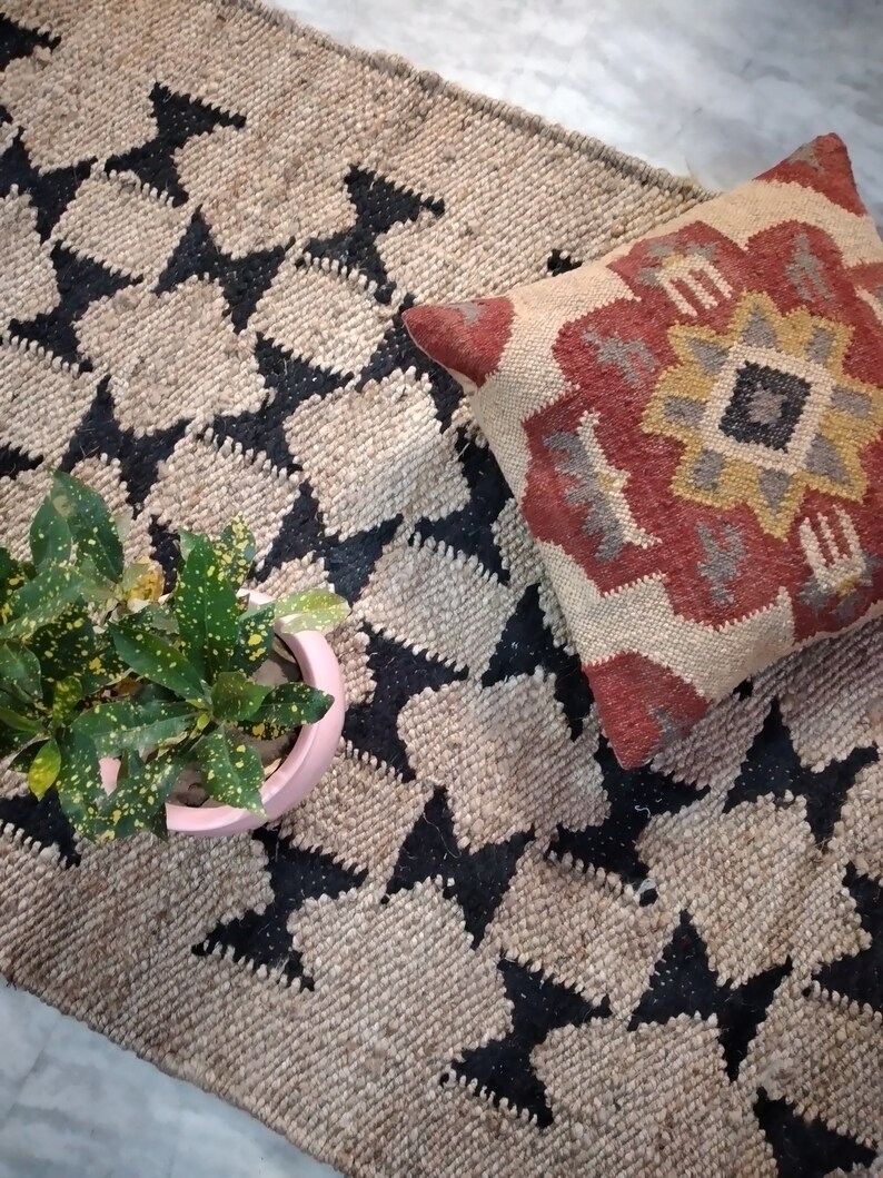 May include: A black and beige jute rug with a geometric pattern. A red, yellow, and gray patterned pillow is on top of the rug.