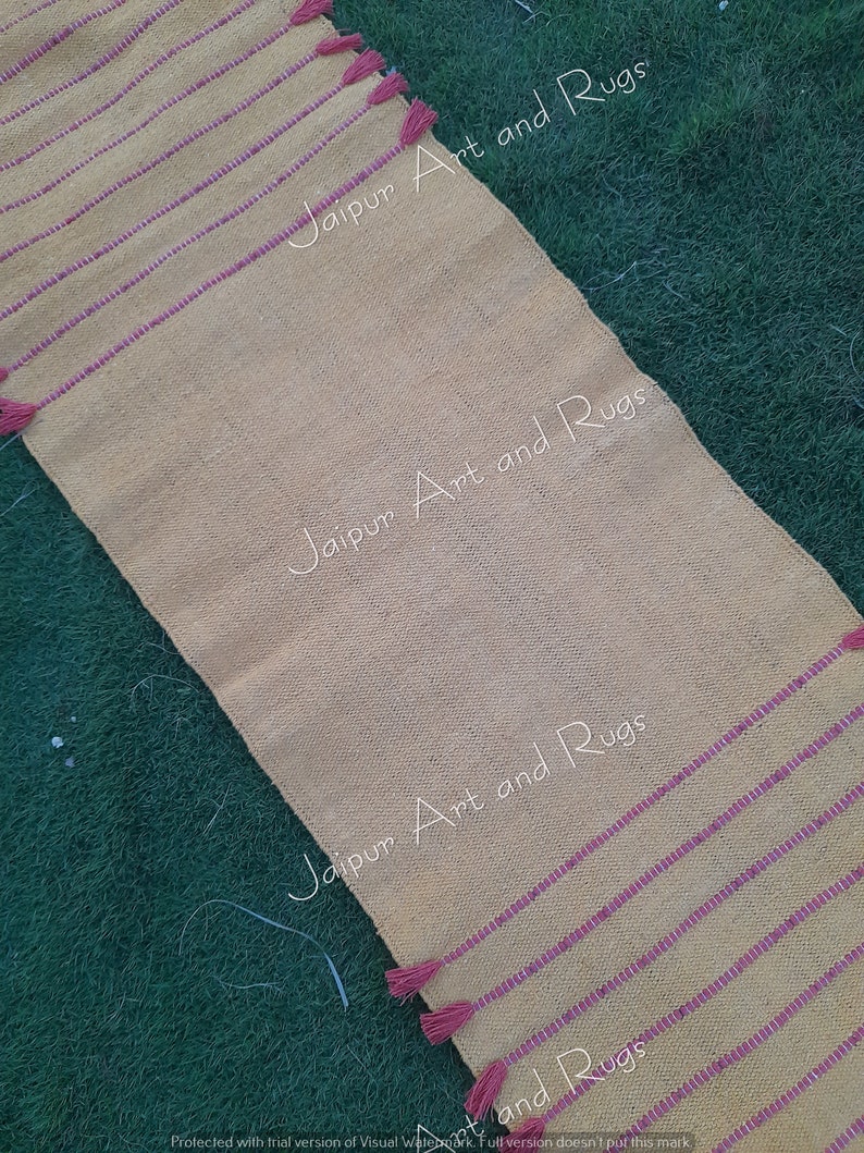 May include: A yellow and pink striped runner rug with tassels on the ends. The rug has the text "Jaipur Art and Rugs" printed on it.