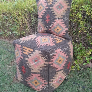May include: A square, gray, patterned ottoman with a matching patterned pillow. The ottoman and pillow are made of woven fabric with pink, yellow, and green accents. The ottoman is sitting on a green grassy surface.
