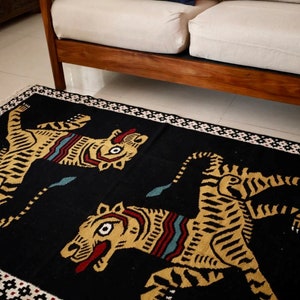 Puede incluir: Alfombra negra con borde blanco y dos tigres amarillos con detalles rojos y azules. La alfombra est&aacute; sobre un suelo blanco delante de un sof&aacute; de madera.