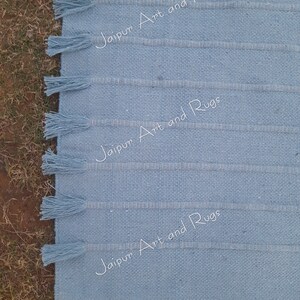 May include: A light blue woven rug with fringe on the edges. The rug has a subtle striped pattern and the text "Jaipur Art and Rugs" is repeated three times on the rug.