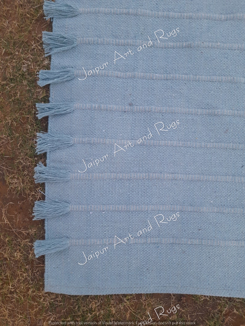 May include: A light blue woven rug with fringe on the edges. The rug has a subtle striped pattern and the text "Jaipur Art and Rugs" is repeated three times on the rug.