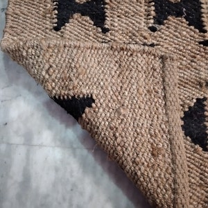 May include: A close-up of a jute rug with a black geometric pattern. The rug is woven with natural fibers and has a textured surface.