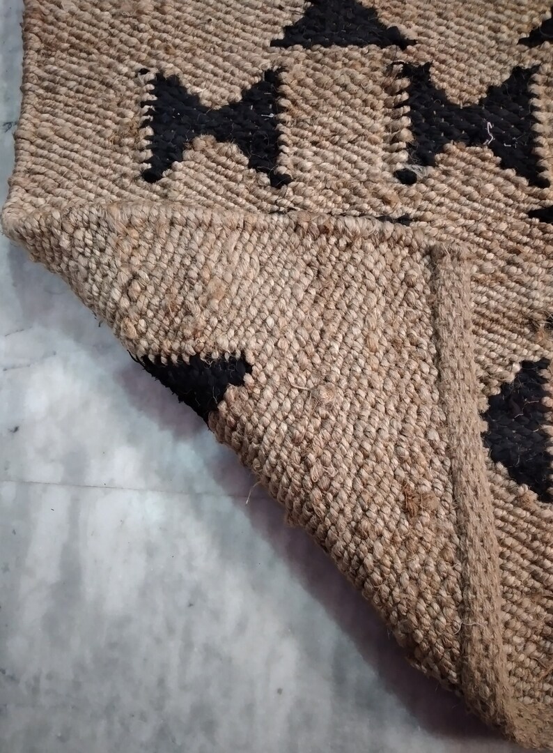 May include: A close-up of a jute rug with a black geometric pattern. The rug is woven with natural fibers and has a textured surface.