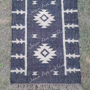 May include: A black and white geometric patterned rug with fringe. The rug has a dark blue background with a white geometric pattern. The rug is made of cotton and is perfect for adding a touch of style to any room.