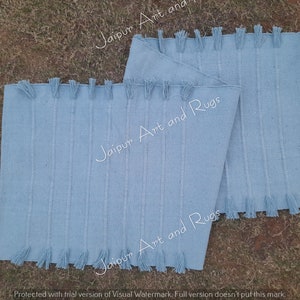 May include: Two light blue cotton rugs with fringe edges. The rugs are stacked on top of each other. The text "Jaipur Art and Rugs" is printed on the rugs.