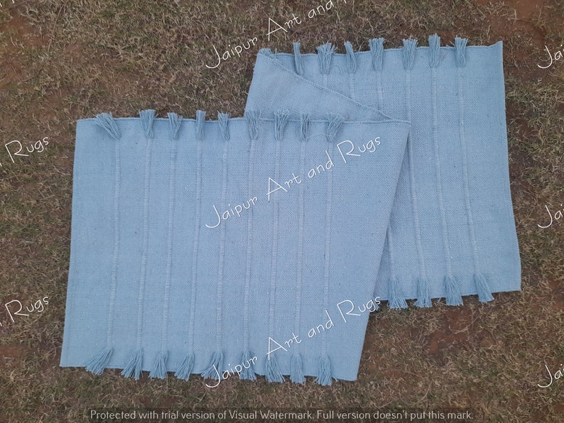 May include: Two light blue cotton rugs with fringe edges. The rugs are stacked on top of each other. The text "Jaipur Art and Rugs" is printed on the rugs.