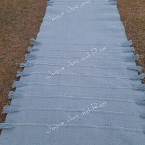 May include: A light blue woven rug with fringe on the edges. The rug has the text "Jaipur Art and Rugs" printed on it three times.