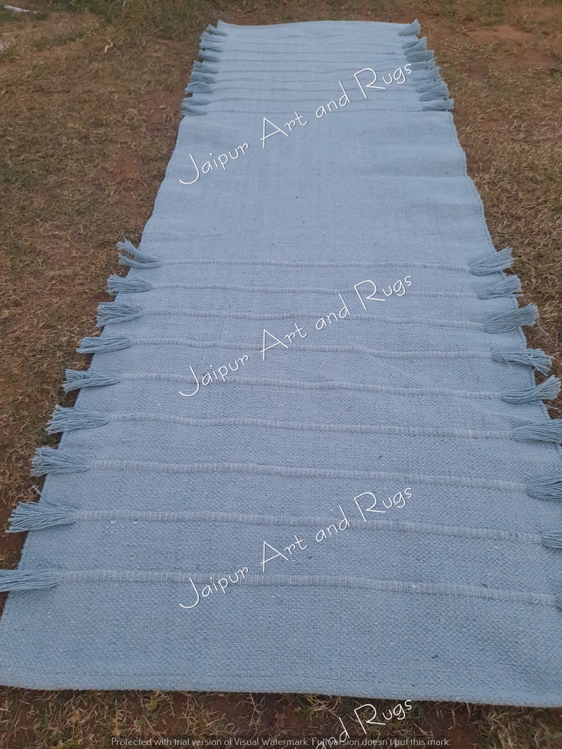 May include: A light blue woven rug with fringe on the edges. The rug has the text "Jaipur Art and Rugs" printed on it three times.