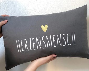 May include: Grey pillow with the text "HERZENSMENSH" in white lettering and a gold heart in the centre.