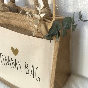 Op de afbeelding: Een natuurlijke jute tas met een witte canvas inzet. De canvas inzet heeft een gouden hart en de tekst "Mommy Bag" in zwart gedrukt.