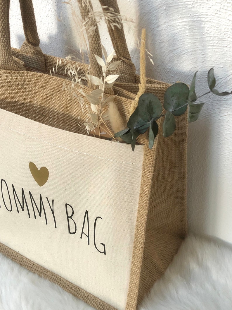 Op de afbeelding: Een natuurlijke jute tas met een witte canvas inzet. De canvas inzet heeft een gouden hart en de tekst "Mommy Bag" in zwart gedrukt.