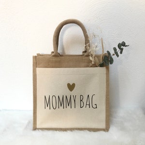 Op de afbeelding: Een beige en bruine draagtas met een witte canvas inzet. De canvas inzet heeft een gouden hart en de tekst "MOMMY BAG" in zwart.