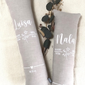 May include: Two light grey, rectangular pillows with white text and floral designs. One pillow reads "Luisa" with birth details: 3600 g, 54 cm. The other reads "Nala" with birth details: 3060 g, 50 cm. Both pillows have "KU 33 cm" text.