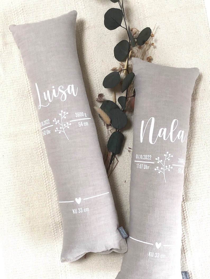 May include: Two light grey, rectangular pillows with white text and floral designs. One pillow reads "Luisa" with birth details: 3600 g, 54 cm. The other reads "Nala" with birth details: 3060 g, 50 cm. Both pillows have "KU 33 cm" text.