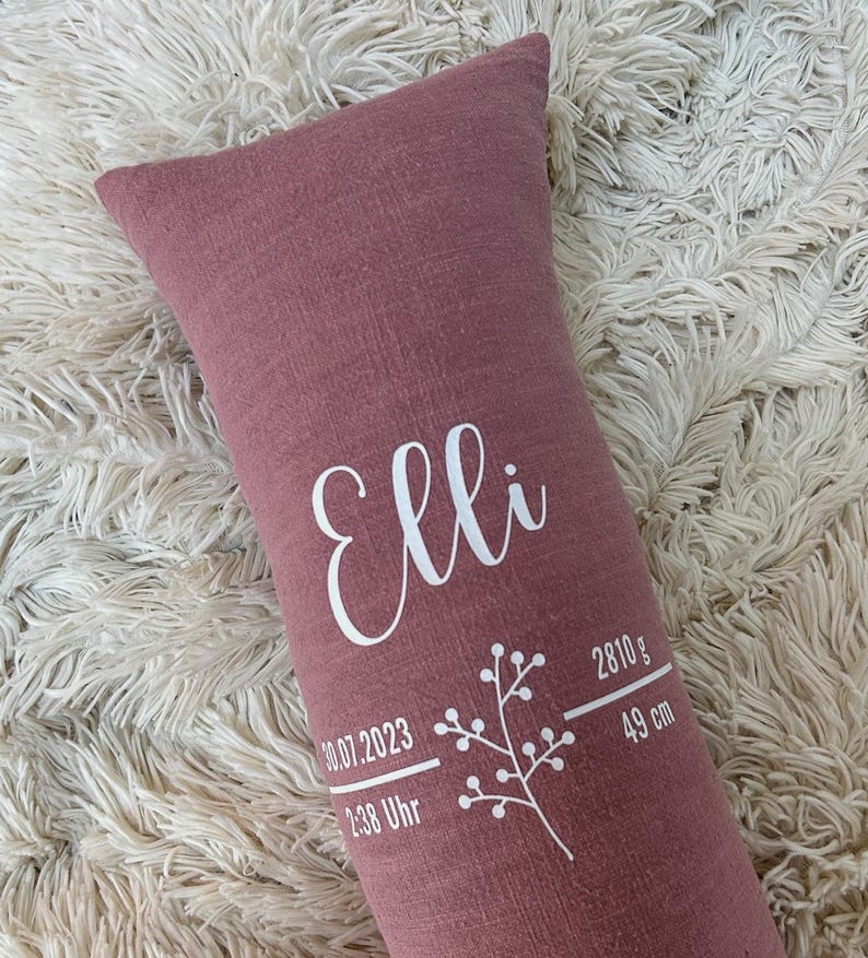 May include: A dusty rose rectangular pillow with the name "Elli" in white script. The pillow also features the birth date, time, weight (2810 g), and length (49 cm) in white text, along with a floral design.
