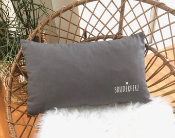 May include: Grey rectangular cushion with white embroidery that reads "BRUDERHERZ" with a small heart symbol above the text. The cushion is sitting on a white fluffy surface.