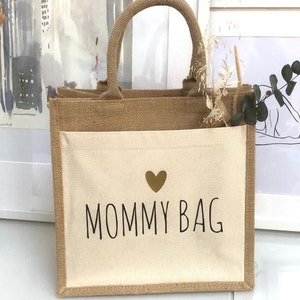 Op de afbeelding: Een bruine jute draagtas met een cr&egrave;mekleurige canvas zak. De zak heeft een gouden hart en de tekst "MOMMY BAG" in zwart.