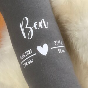 May include: A grey fabric item with the name "Ben" in white script. Below, a white heart and details: "11.09.2023", "7:28 Uhr", "3244 g", and "52 cm". The item is resting on a white, fluffy surface.