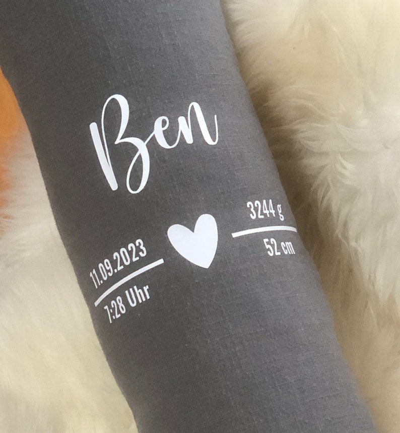 May include: A grey fabric item with the name "Ben" in white script. Below, a white heart and details: "11.09.2023", "7:28 Uhr", "3244 g", and "52 cm". The item is resting on a white, fluffy surface.