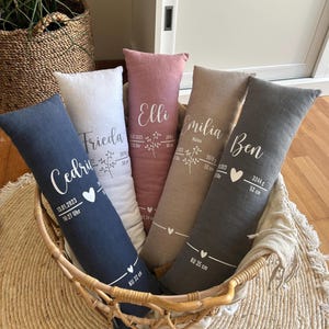 May include: A selection of personalised, long, rectangular pillows in a wicker basket. Each pillow is a different colour, including blue, white, pink, tan, and grey. The pillows have names and birth details printed on them, with a heart symbol.