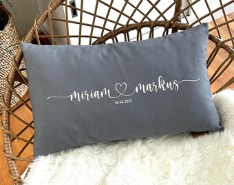 Könnte beinhalten: Ein graues rechteckiges Kissen mit weißem Schriftzug "miriam & markus", einem Herzsymbol zwischen den Namen und dem Datum "06.06.2025". Das Kissen liegt auf einer weißen, flauschigen Oberfläche.