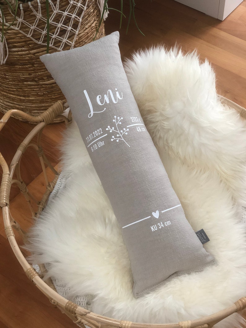 May include: A grey rectangular pillow with white text and a floral design. The text includes the name "Leni", date, time, and measurements. The pillow is resting on a white fluffy surface.