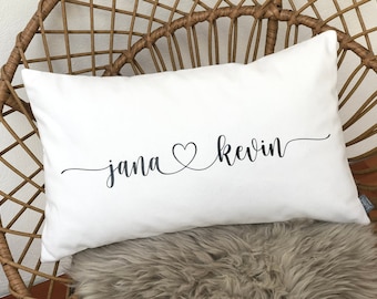 Könnte beinhalten: Weißes rechteckiges Kissen mit den Namen "jana" und "kevin" in schwarzer Schrift, verbunden durch ein Herz. Das Kissen liegt auf einem Korbstuhl mit einem Kunstfell.