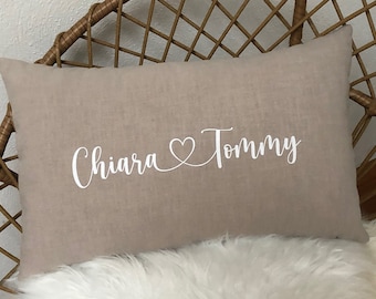 Könnte beinhalten: Ein beigefarbenes Leinenkissen mit den Namen "Chiara" und "Tommy", die in Weiß mit einem Herzsymbol dazwischen gestickt sind.