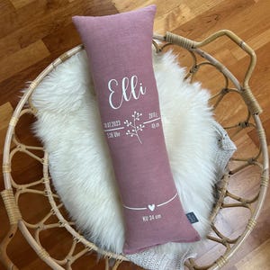 May include: A dusty rose-coloured rectangular pillow with the name "Elli" in white script. The pillow features a floral design, date, time, weight, and length in white. The pillow is 49 cm long and 34 cm in circumference.