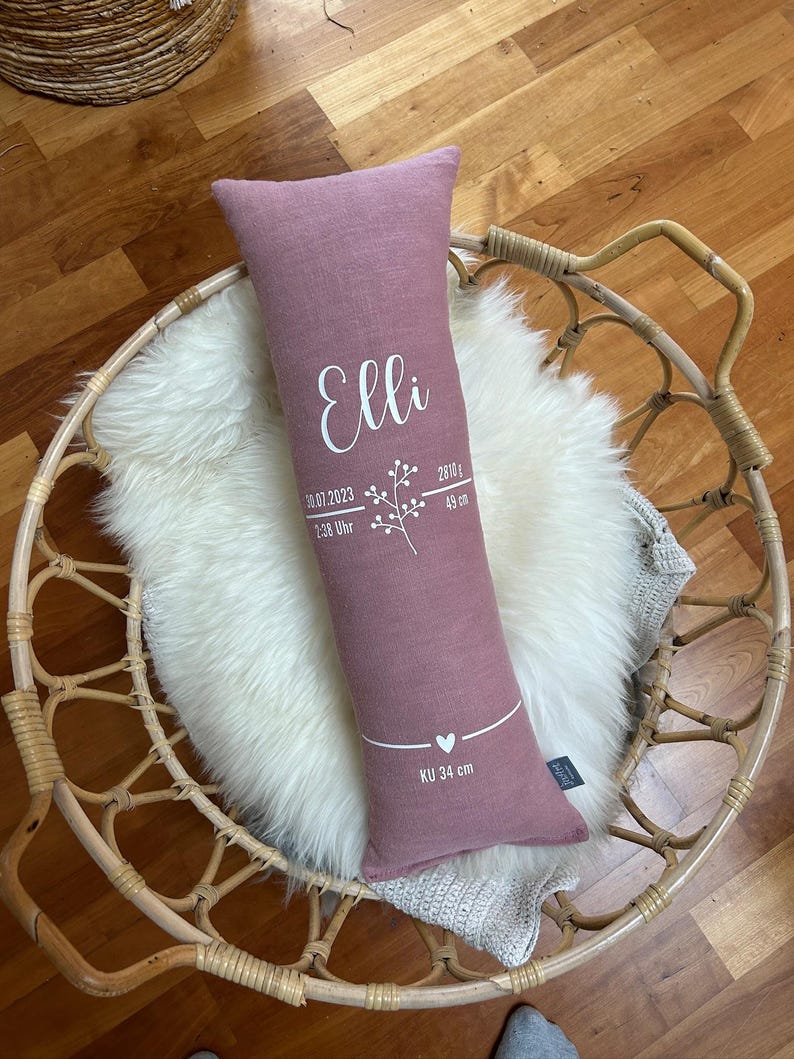 May include: A dusty rose-coloured rectangular pillow with the name "Elli" in white script. The pillow features a floral design, date, time, weight, and length in white. The pillow is 49 cm long and 34 cm in circumference.