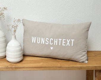 May include: A rectangular beige throw pillow featuring the white text WUNSCHTEXT printed in the centre, accompanied by a small white heart icon below the text. The pillow is positioned next to two white ceramic vases containing dried flowers.