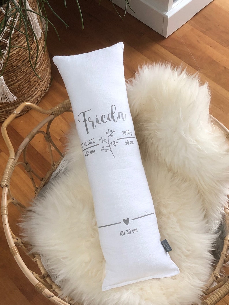 May include: A white rectangular pillow with the name "Frieda" printed in silver script, along with birth details. The pillow is 50 cm long and weighs 3070 grams. It rests on a fluffy white sheepskin rug.